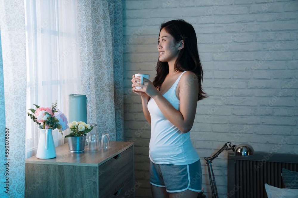 side fresh young asian woman in sleep wear drinking tea standing by window sill and looking out. Pre