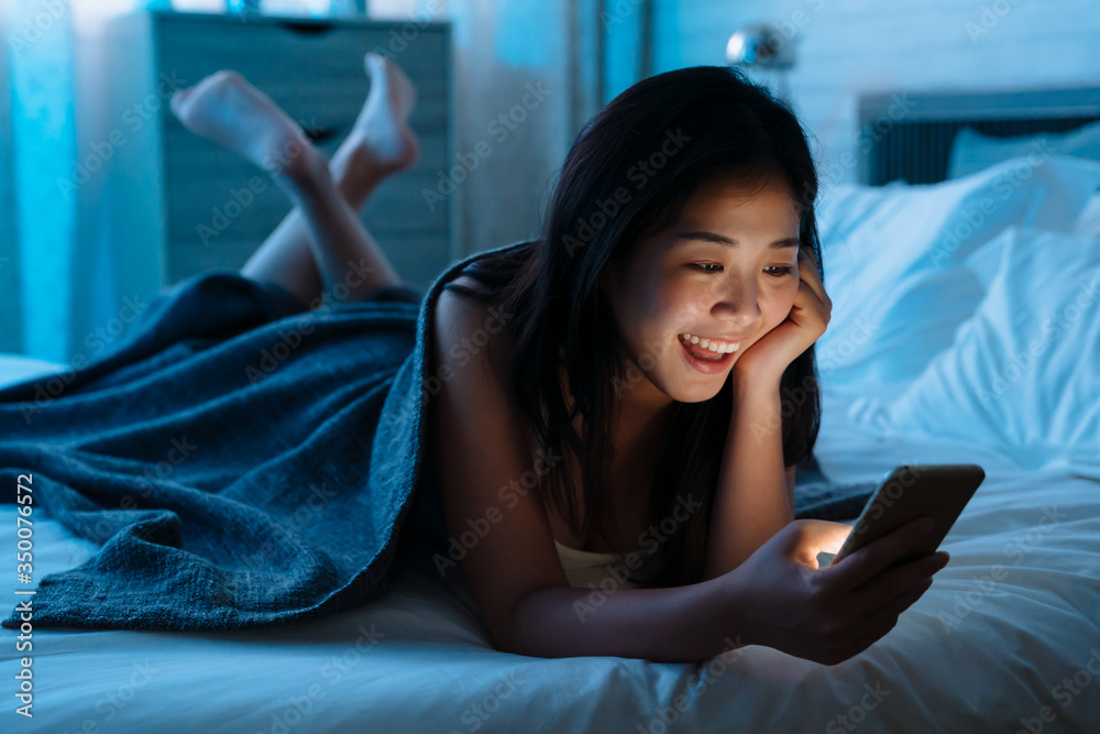 Happy asian japanese teen lying in front in bed at home with smartphone. Young joyful woman watching