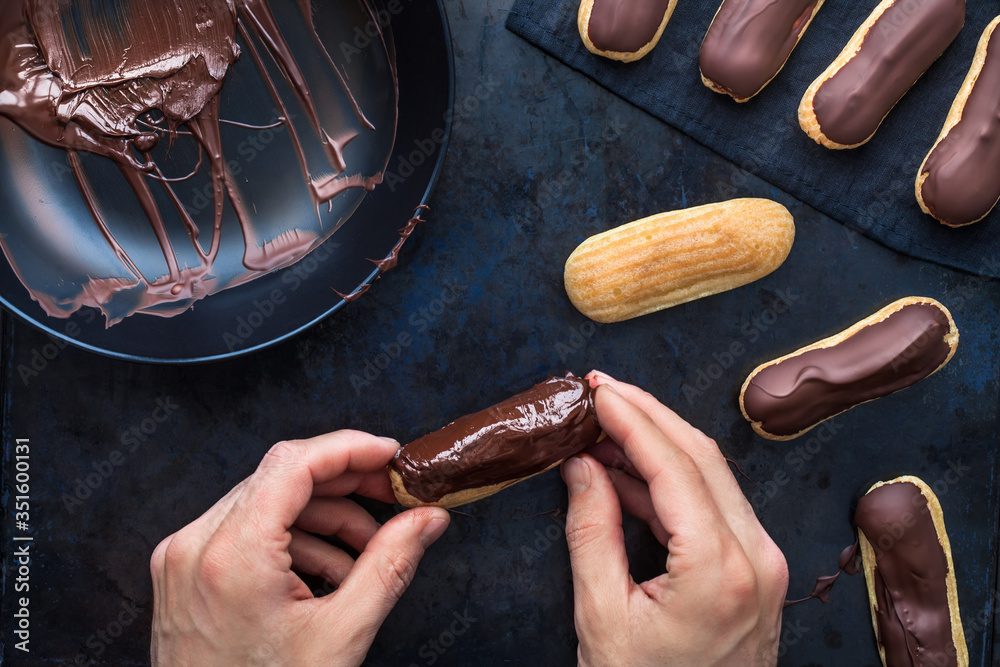 Man prepares eclairs or profiteroles, he covers them with chocolate on an old metal desk dark. Tradi