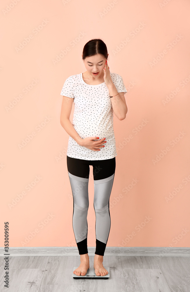 Young pregnant woman measuring her weight against color wall