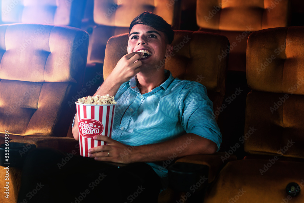 A young man smiling enjoying with his popcorn while watching a movie in theater cinema