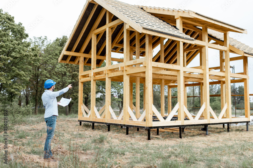 Architect or builder standing with blueprints near the wooden house structure on the construction si