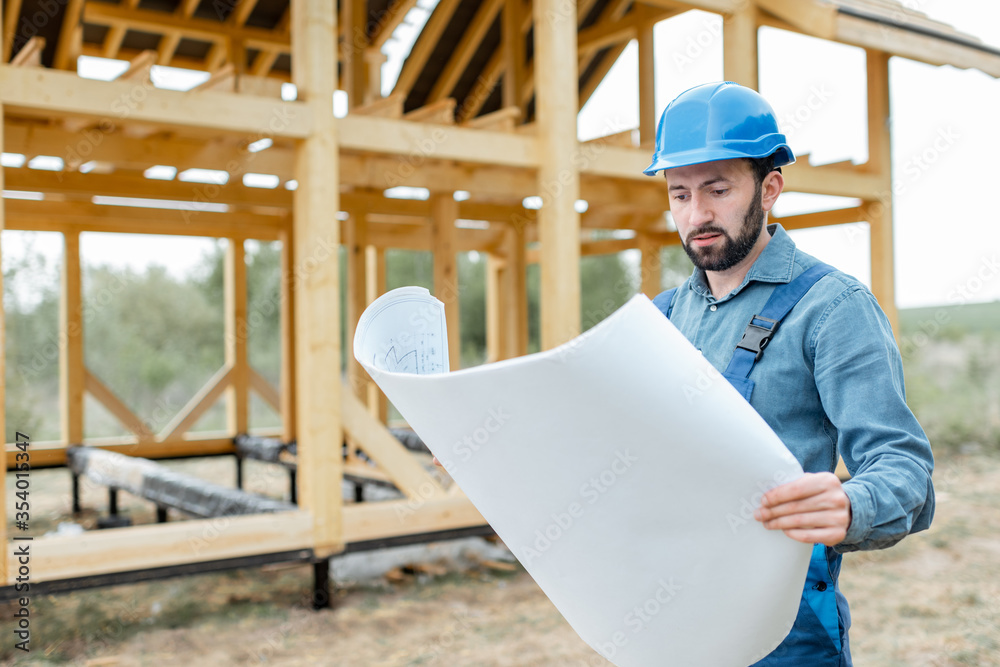 Builder in blue overalls and hard hat with blueprints on the construction site, building wooden fram