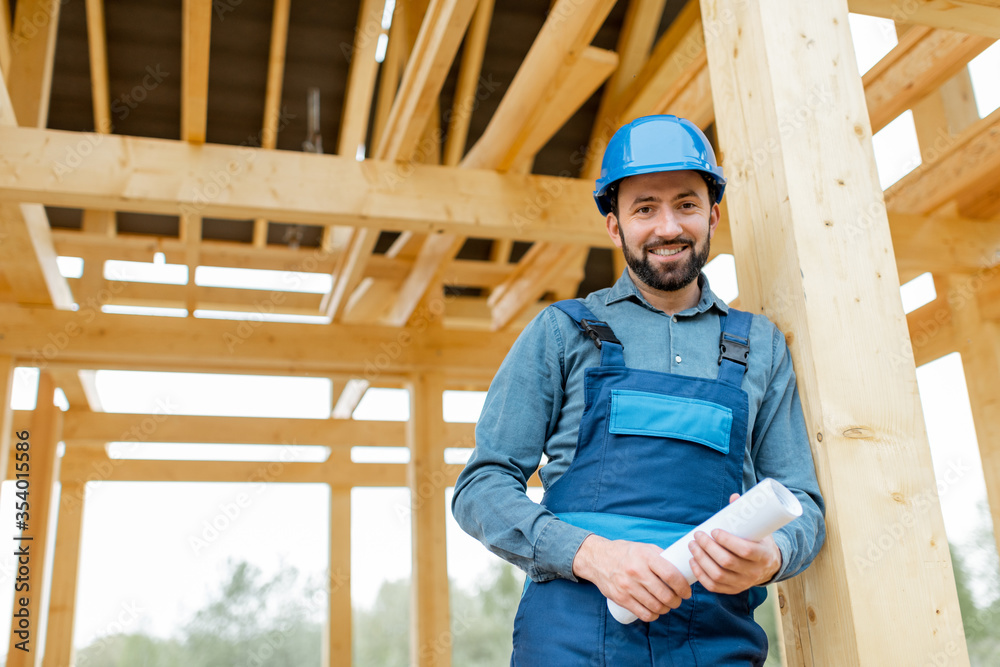 Portrait of a handsome builder in blue overalls and hard hat with blueprints on the construction sit