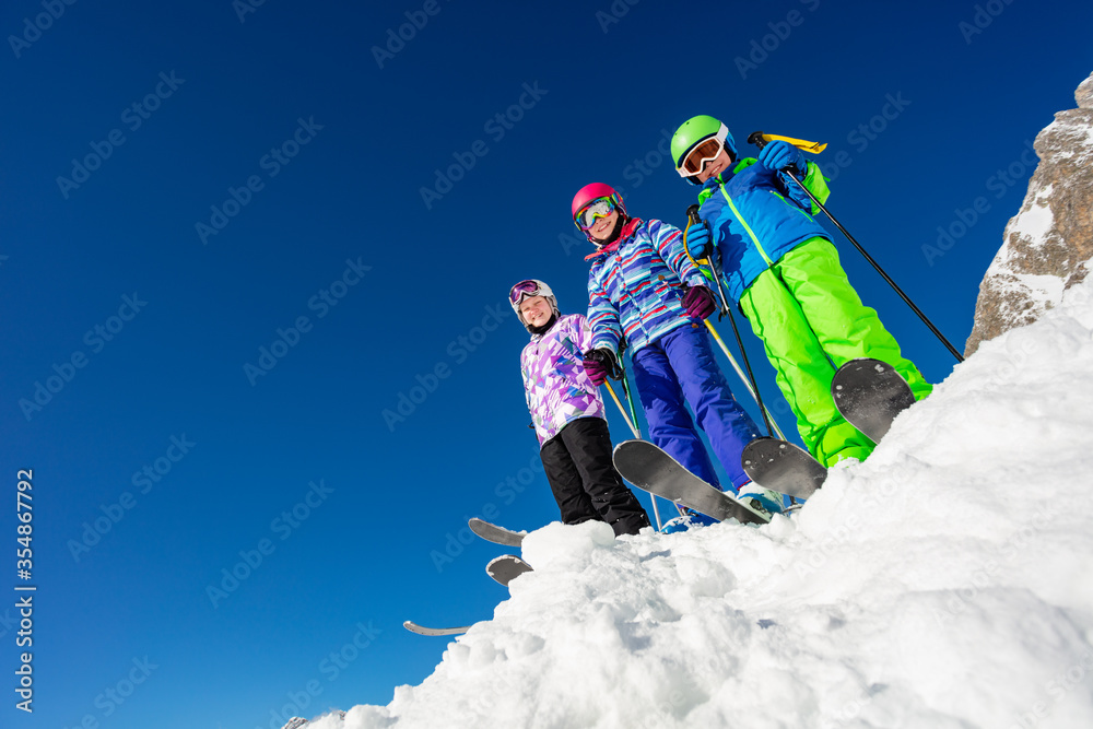 从下面看，三个孩子站在山顶上，穿着五颜六色的滑雪服