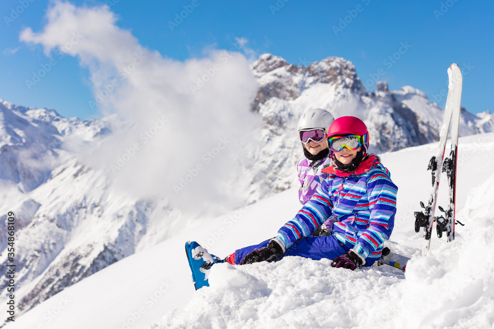 Two happy girls sit in the mountain with ski over high peaks and clouds in snow look at camera smili