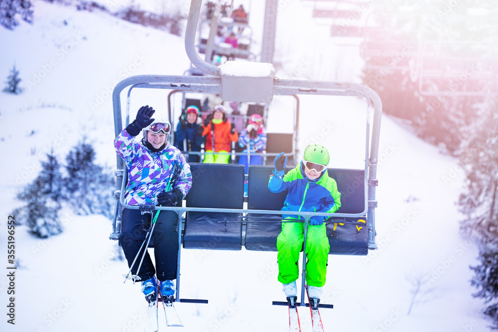 Boy with girl sit on the ski chair lift together and wave hands on Alpine mountain resort