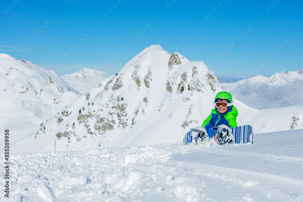 Portrait of a little snowboarder sit in snow with snowboard over mountain in bright blue and green s