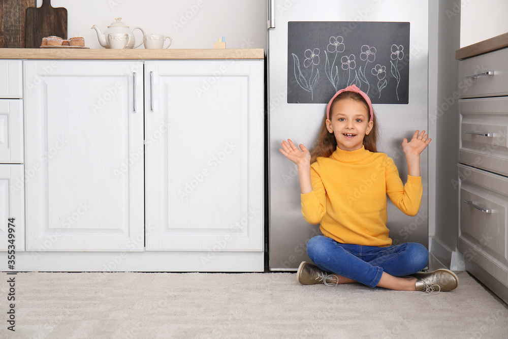 Little girl near chalkboard on refrigerator in kitchen