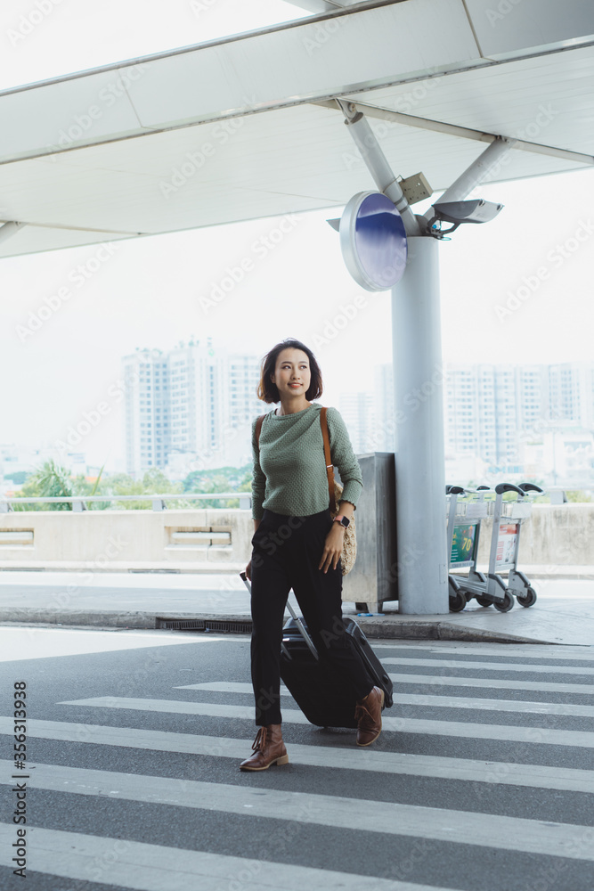 Beautiful woman happily walking along pedestrian strip with suitcase and airport on background