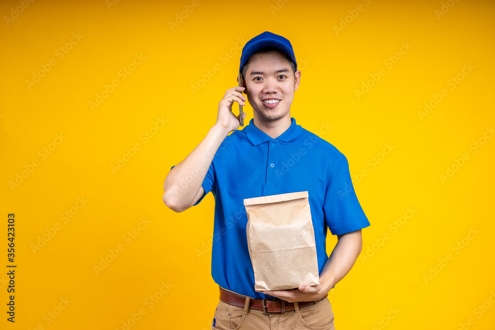 Asian delivery man holding fast food bag and using mobile phone over yellow isolate background. Work