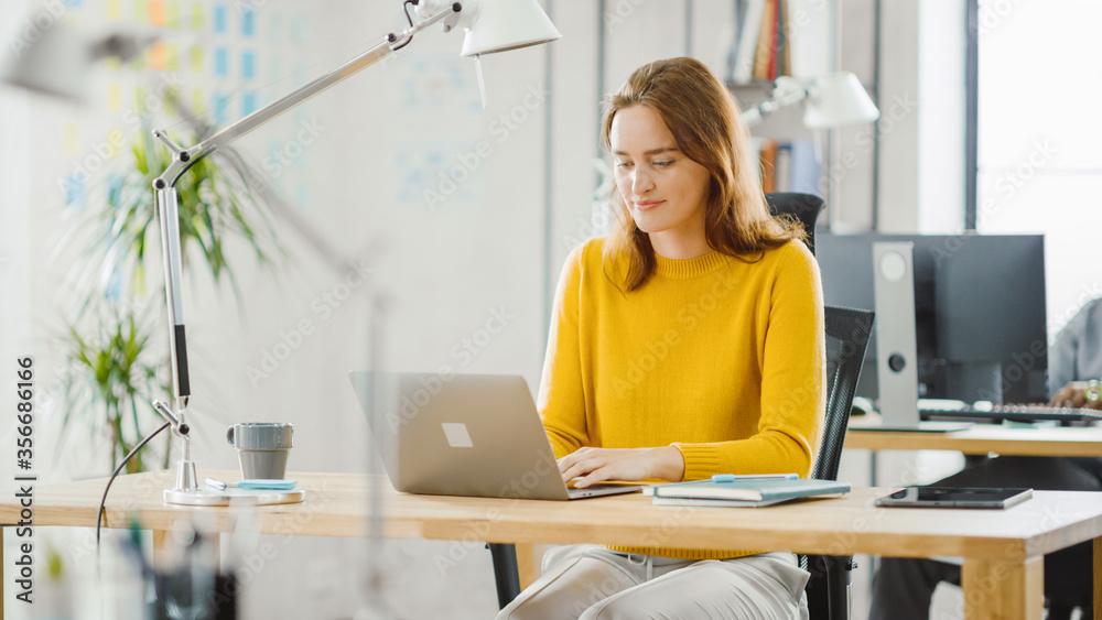 Beautiful and Creative Young Woman Sitting at Her Desk Using Laptop Computer. In the Background Brig