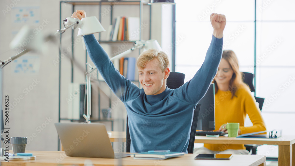 Handsome Caucasian IT Specialist Sitting at His Desk works on a Laptop, Raises Arms in Celebration a