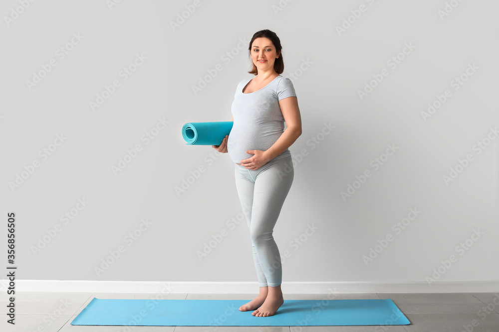 Young pregnant woman with yoga mat near light wall