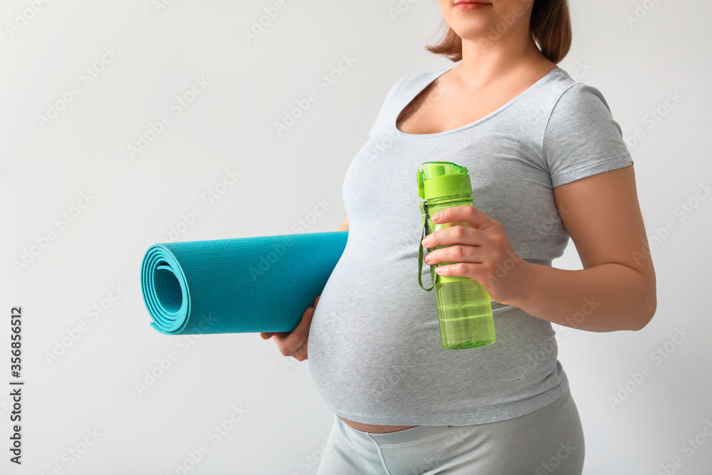 Young pregnant woman with yoga mat and water near light wall