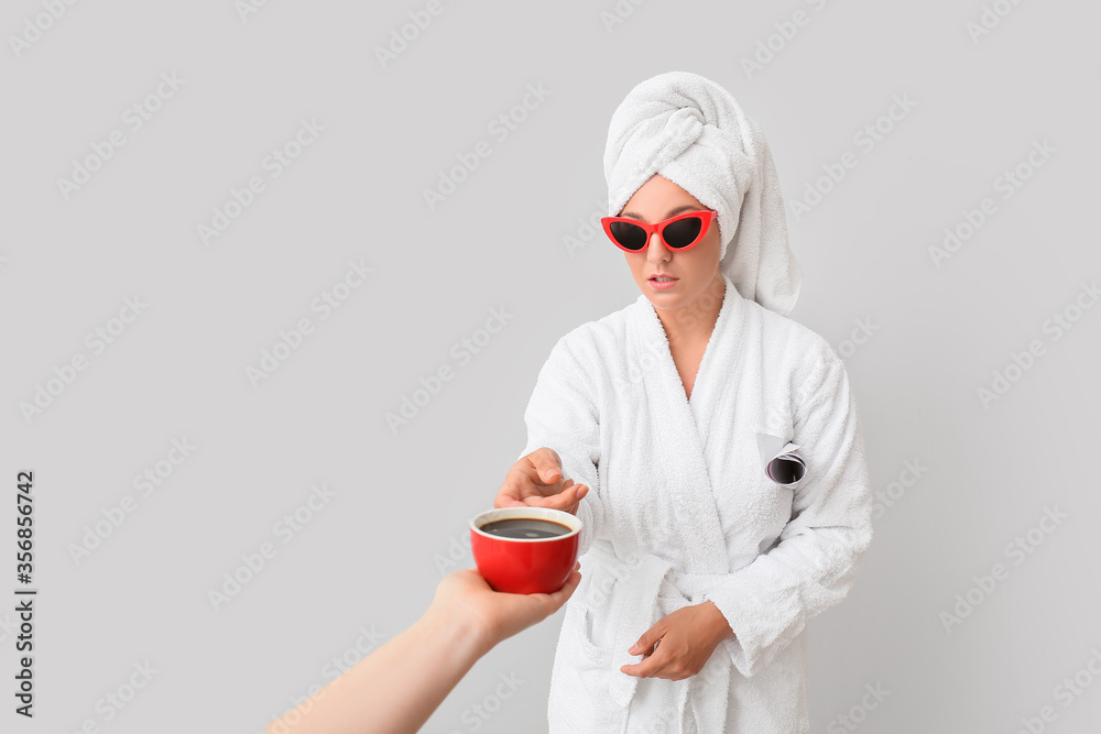 Hand offering cup of coffee to young woman in bathrobe on grey background