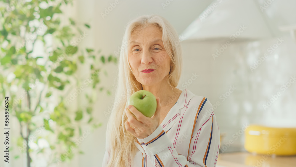 老年女性养老金领取者在家里明亮的厨房里吃苹果的真实肖像。太美了