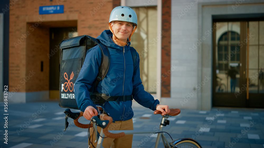 Handsome Happy Food Delivery Man Wearing Thermal Backpack and Safety Helmet Stands Beside his Bike i