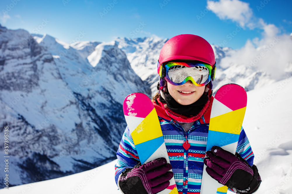 Close-up portrait of cute girl in pink helmet and color hold crossed pair of ski smiling, goggles ov