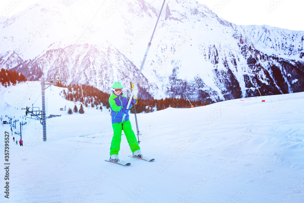 Happy little boy lifting on the ski drag lift rope in bright sport outfit over sunset light near mou