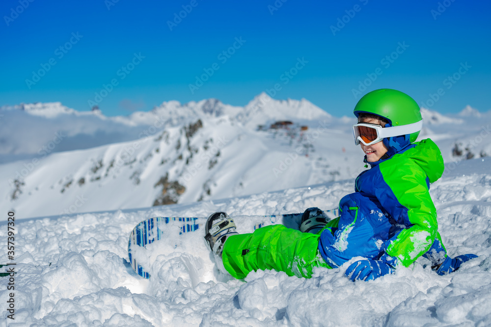 一个男孩穿着亮蓝色和绿色的运动服坐在雪地里，滑雪板翻山的肖像