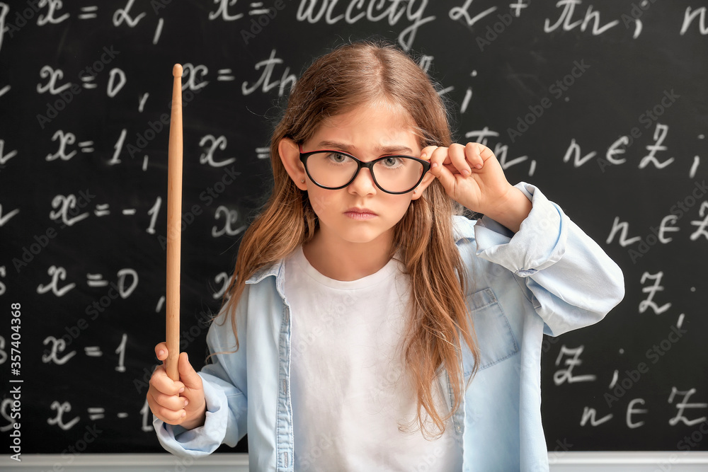 Cute little schoolgirl with pointer near blackboard in classroom