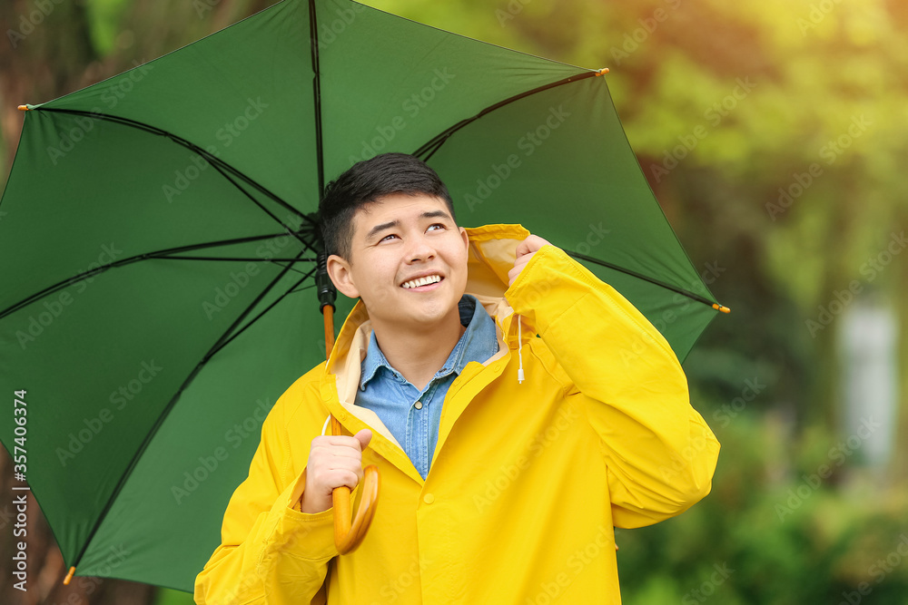 亚洲年轻男子在公园里打着雨伞，穿着雨衣