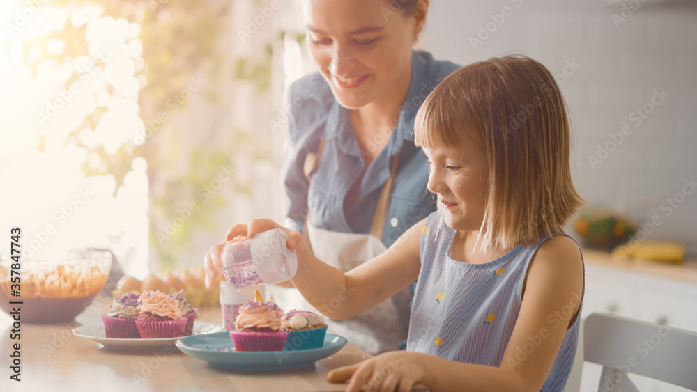 In the Kitchen: Mother and Cute Little Daughter Sprinkling Funfetti on Creamy Cupcakes Frosting. The