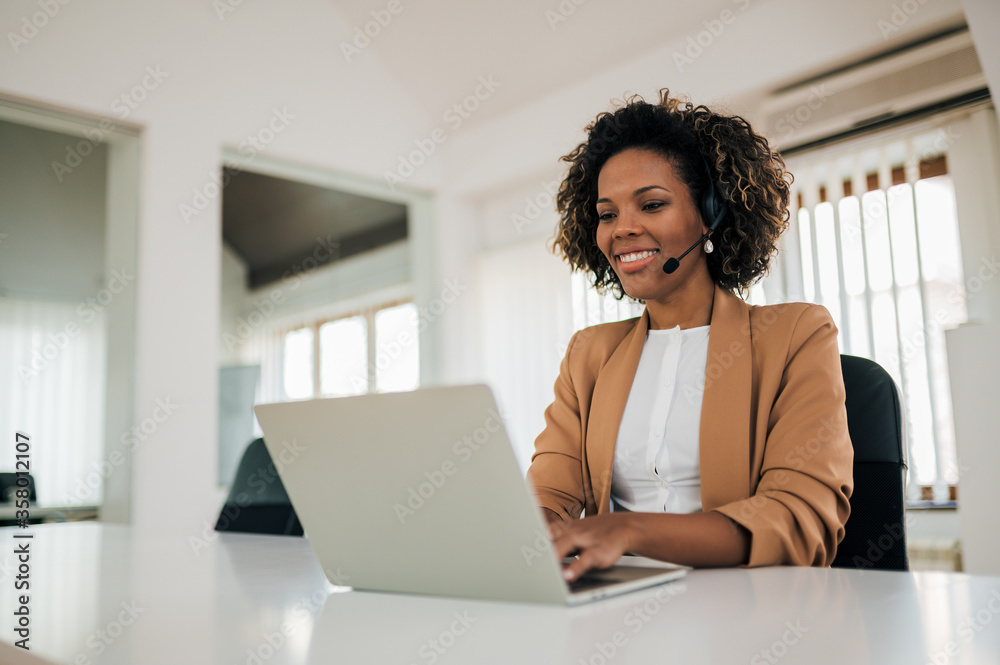 Portrait of a positive mixed race secretary working on laptop, wearing headset.