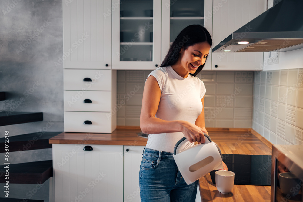 Portrait of a smiling woman pouring hot water in a cup while standing at the kitchen.