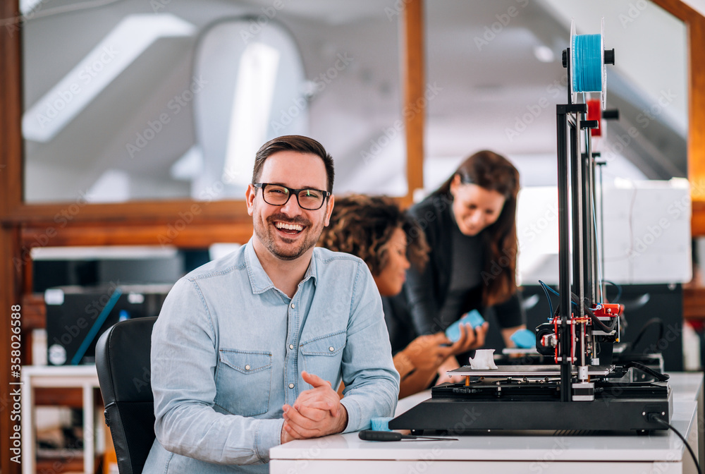 Portrait of a happy engineer with 3D printer, looking at camera.