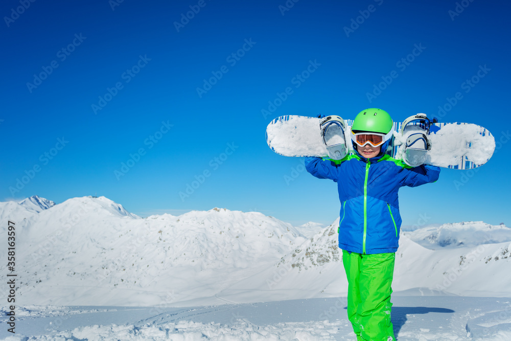 可爱的男孩站在雪地里，把滑雪板放在肩膀上，戴着滑雪帽，在干净的山景上