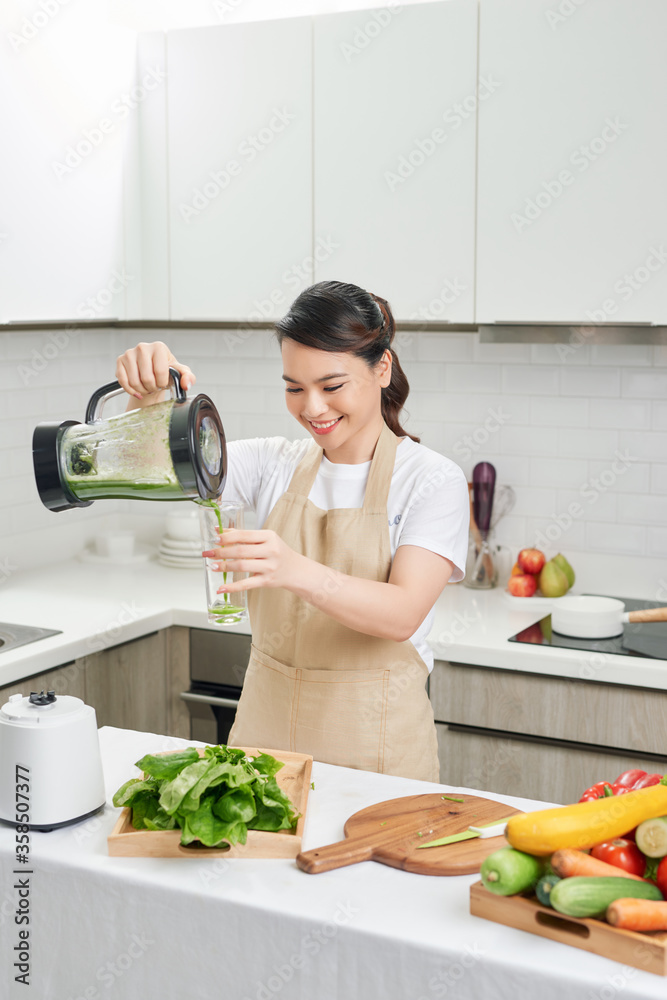 穿着亚麻围裙的年轻女性将绿色奶昔饮料从搅拌机倒到装满蔬菜的瓶子里