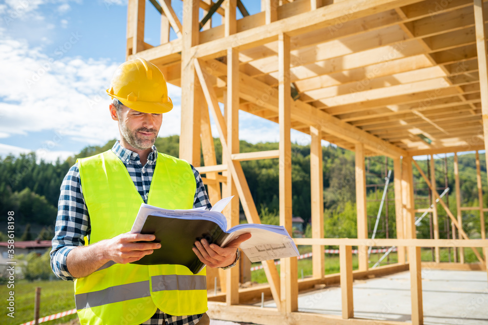 Engineer with hardhat and blueprints on building site of wood frame house under construction