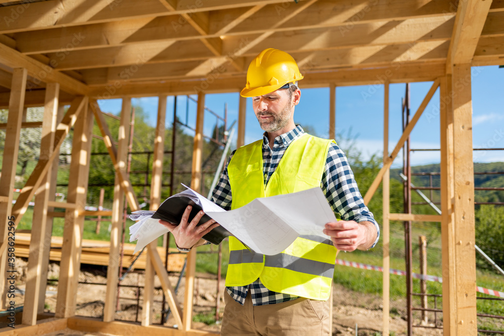 Construction engineer or architect with blueprints visiting building site of wood frame house