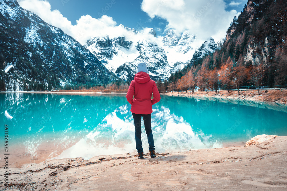 Young woman in red jacket is standing on shore of lake with azure water in autumn in Dolomites, Ital