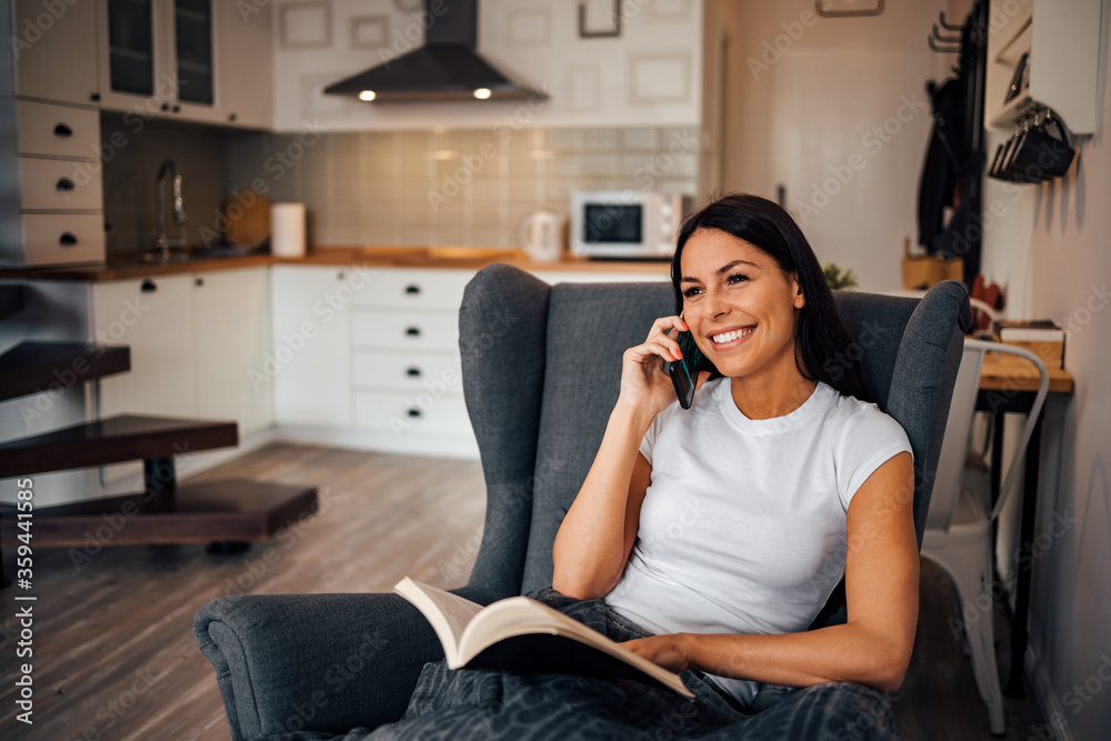 Happy woman relaxing at home, portrait. Talking on smart phone while reading a book.