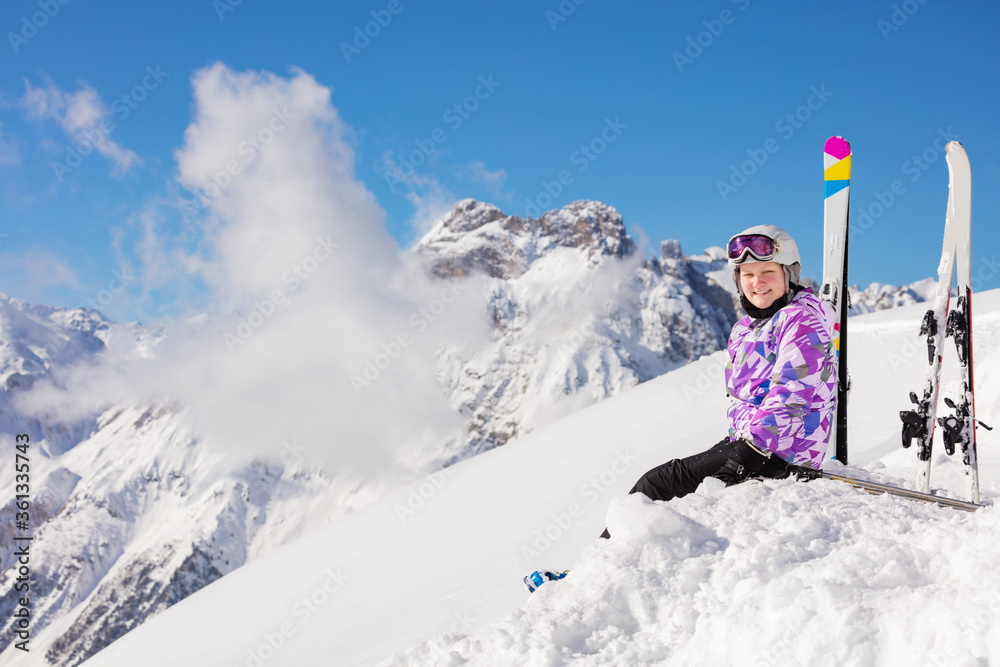 快乐的女孩，穿着滑雪服，摘下头盔和口罩，坐在山顶的雪地里看着镜头