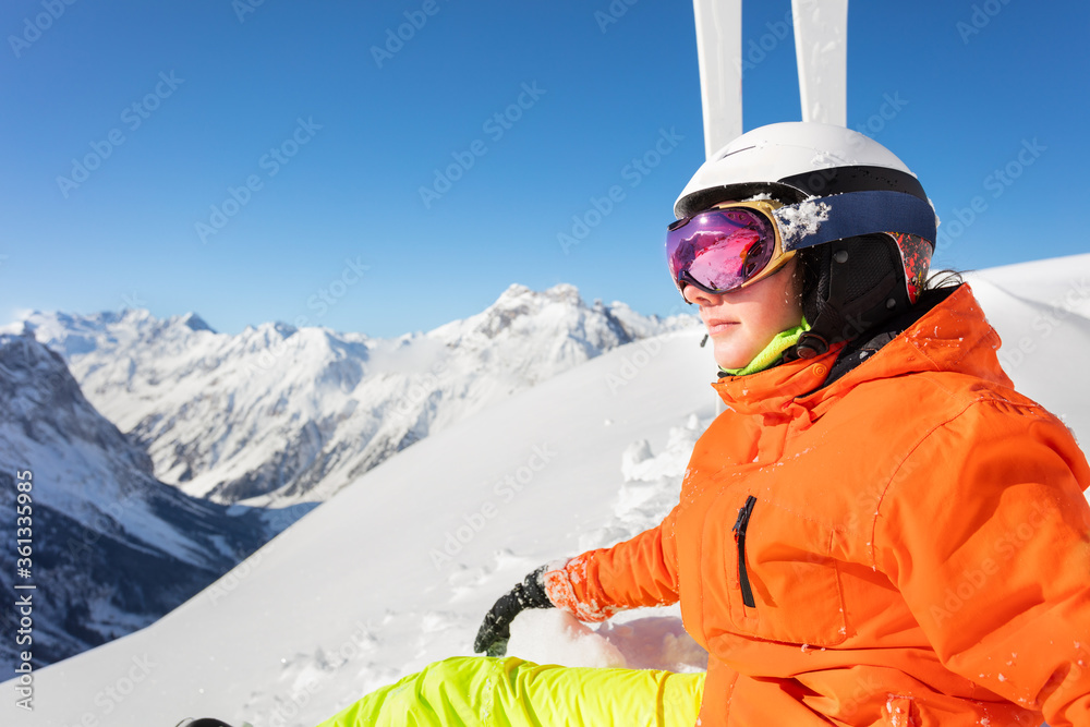 Close portrait with mask on of a teenage girl in orange outfit near ski and look on the mountain