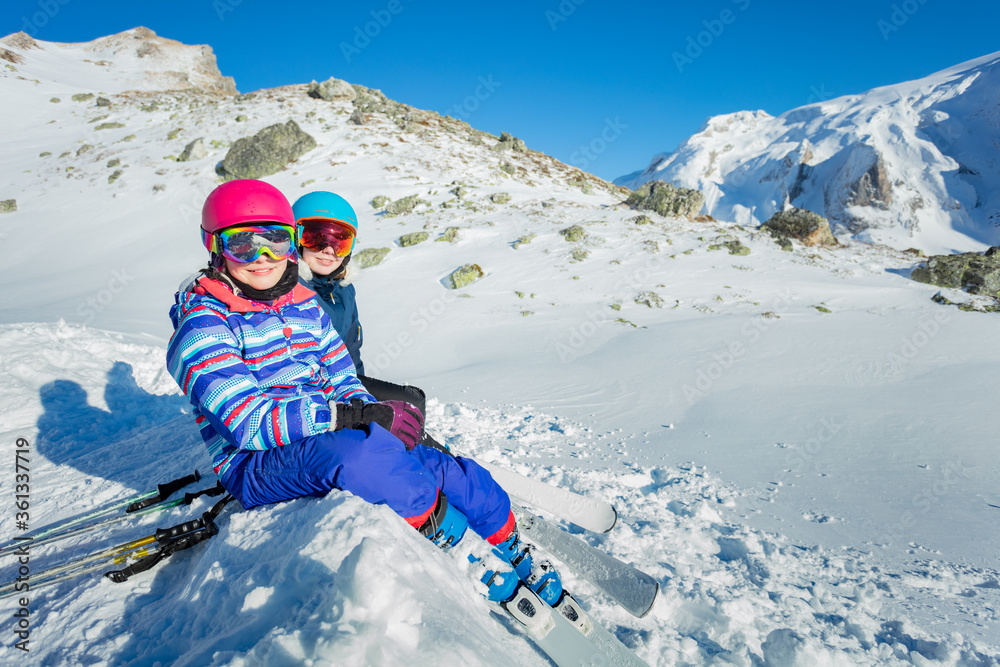 两个快乐的女孩戴着滑雪面罩和头盔坐在雪地上看山