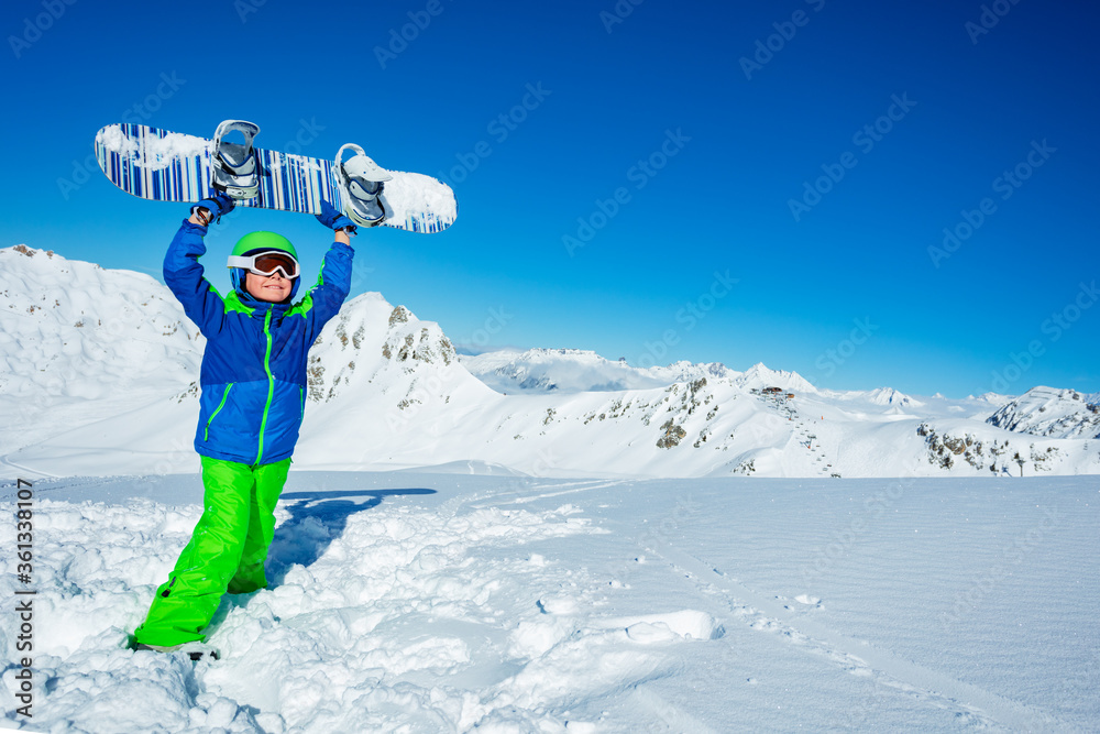 小男孩戴着面罩，戴着滑雪头盔，站在雪地上，双手将滑雪板举过头顶