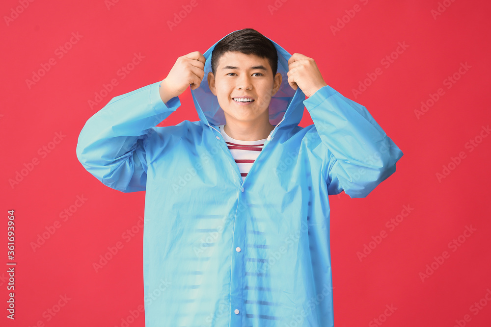 Young Asian man in raincoat on color background