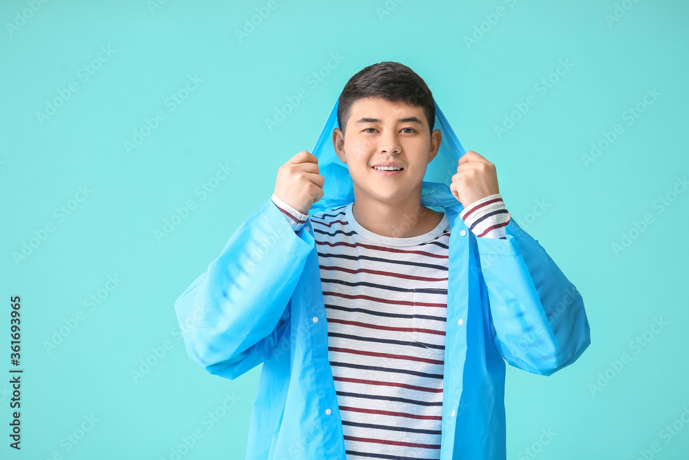 Young Asian man in raincoat on color background