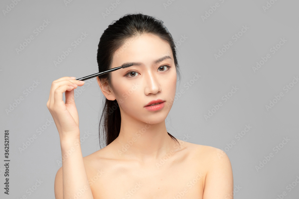 灰色背景下使用眉笔的亚洲美女特写脸，化妆护肤