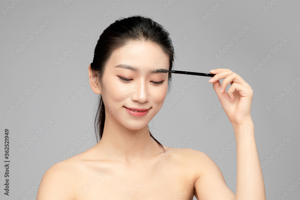 灰色背景下使用眉笔的亚洲美女特写脸，化妆护肤