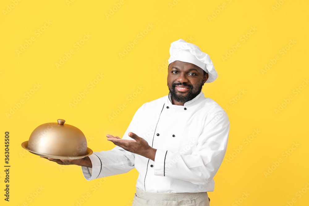 Male African-American chef with tray and cloche on color background