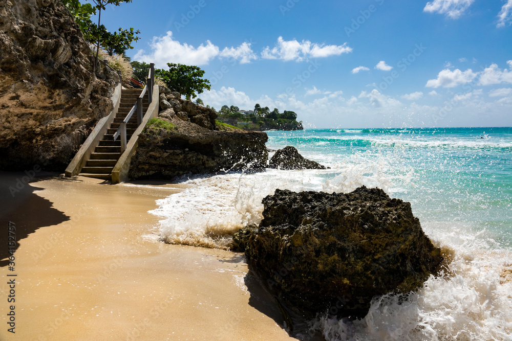 Waves crash into a large rock on the sandy shore and splash high in the air.