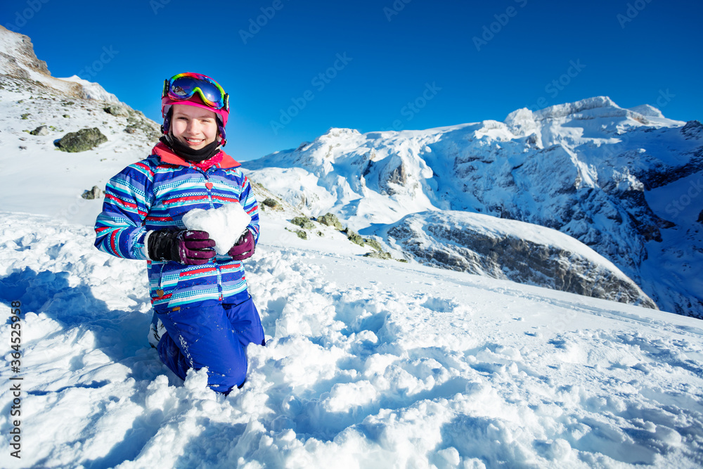 滑雪小女孩抱着雪呈心形微笑，背景是山顶站在她身上