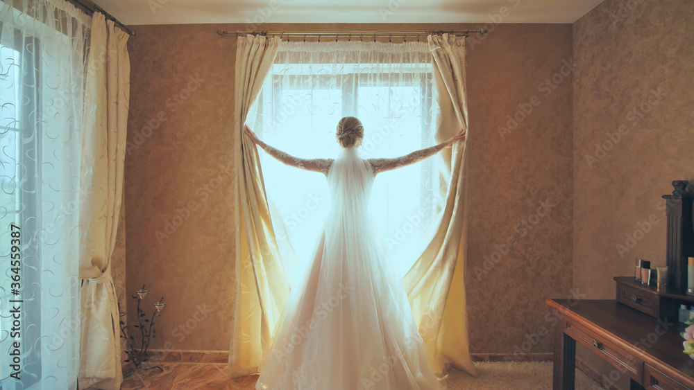 The bride opens the curtains on the window at home.