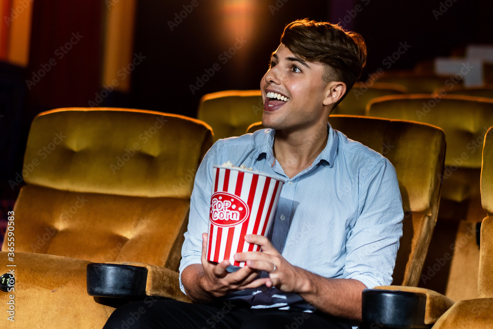 A young man smiling enjoying with his popcorn while watching a movie in theater cinema